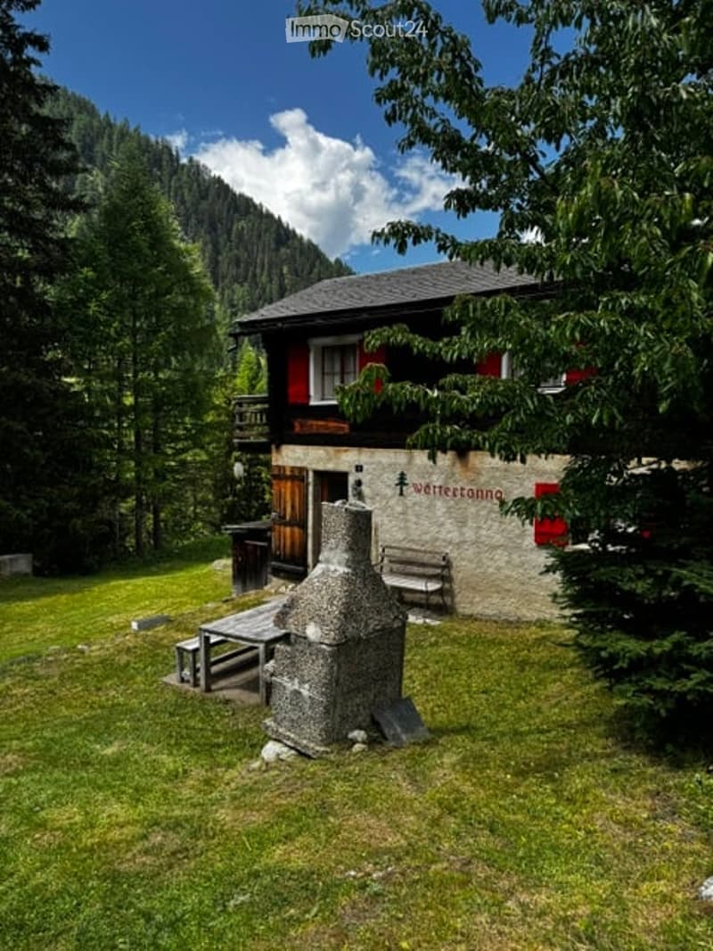 A rustic wooden chalet with red shutters and a stone chimney, surrounded by a grassy area with a wooden bench and table, set against a backdrop of mountains and trees.