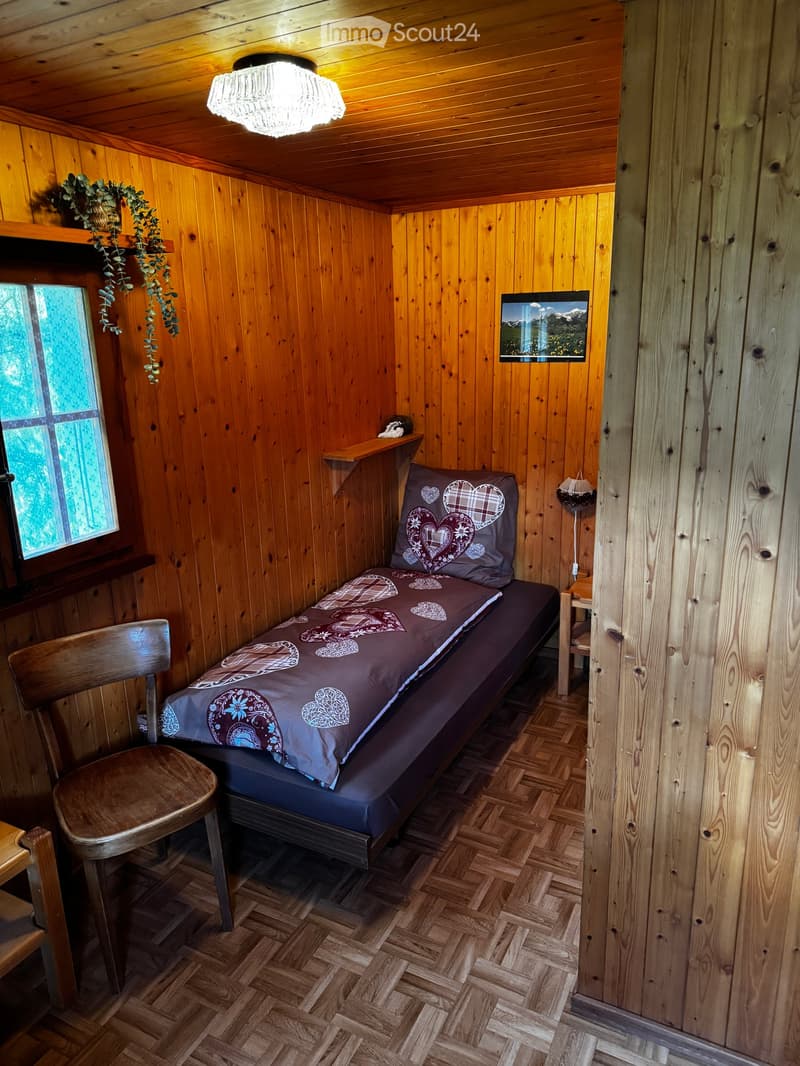 Cozy wooden bedroom with a single bed, patterned bedding, a wooden chair, a small table, and a decorative picture frame on the wall.