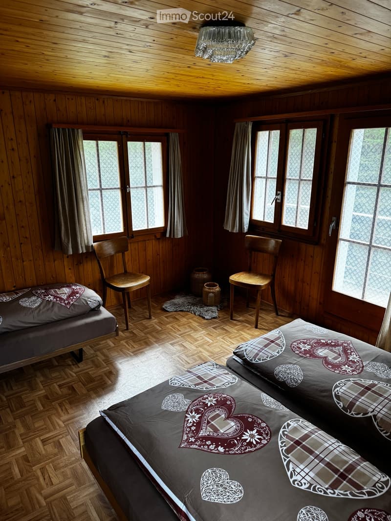 Cozy wooden bedroom with two beds, wooden chairs, large windows with curtains, and a chandelier.