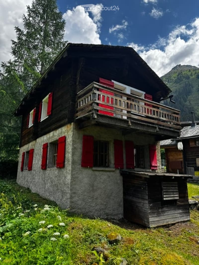 Traditional wooden chalet with red shutters, stone base, and balcony overlooking a mountain view.