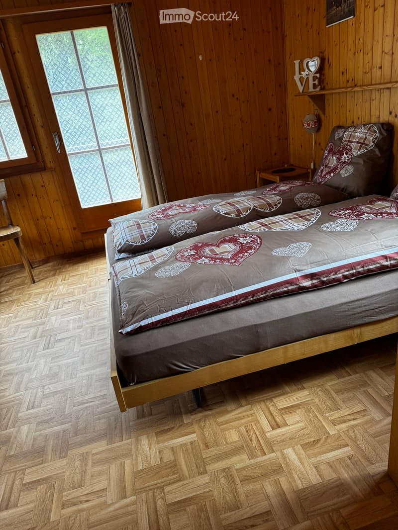 Cozy bedroom with wooden walls and herringbone wooden flooring, featuring a double bed with patterned bedding and a nightstand with a lamp.