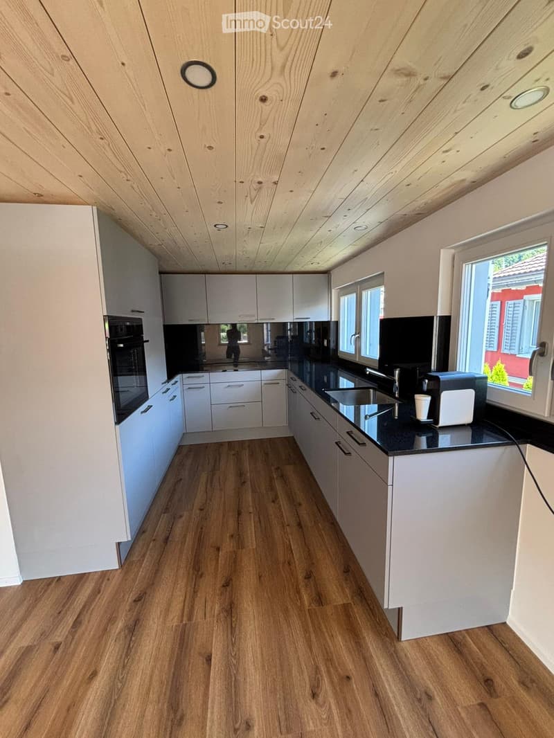 Wooden flooring, white cabinetry, black countertops, large kitchen island, black backsplash, built-in oven, double sink, kitchen faucet, toaster, window with view of a building outside