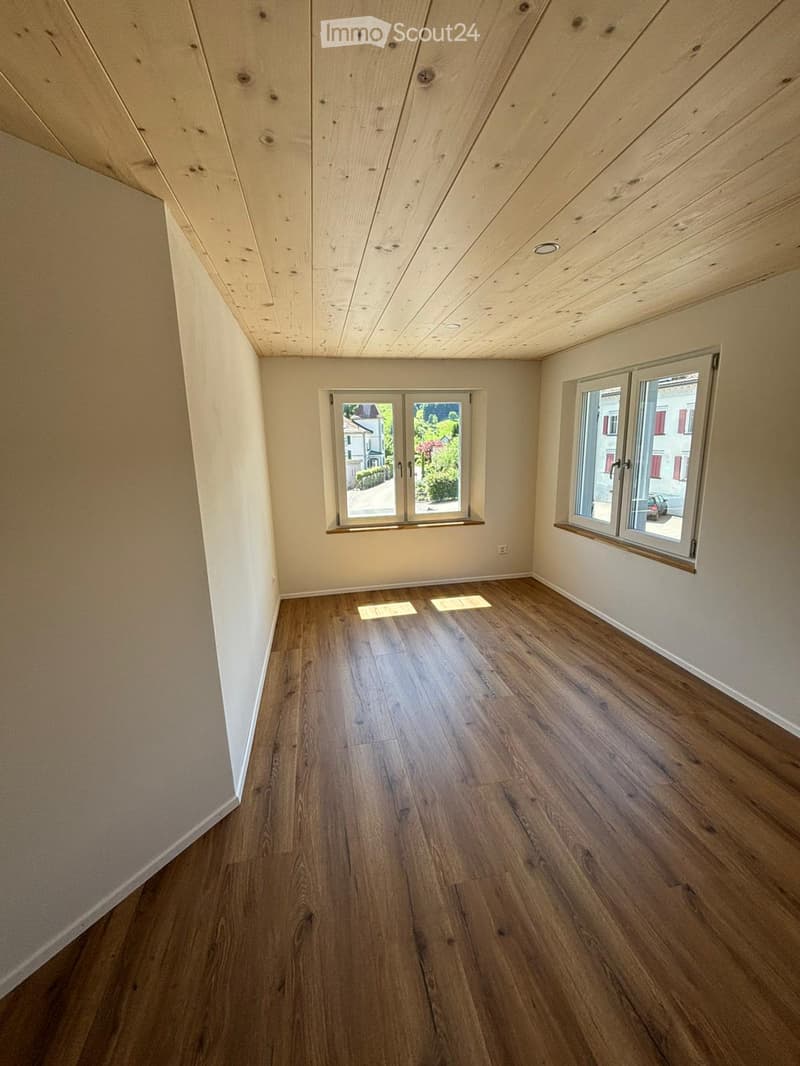 Empty room with wooden floor, wooden ceiling, and white walls. Two large windows on either side of the room.