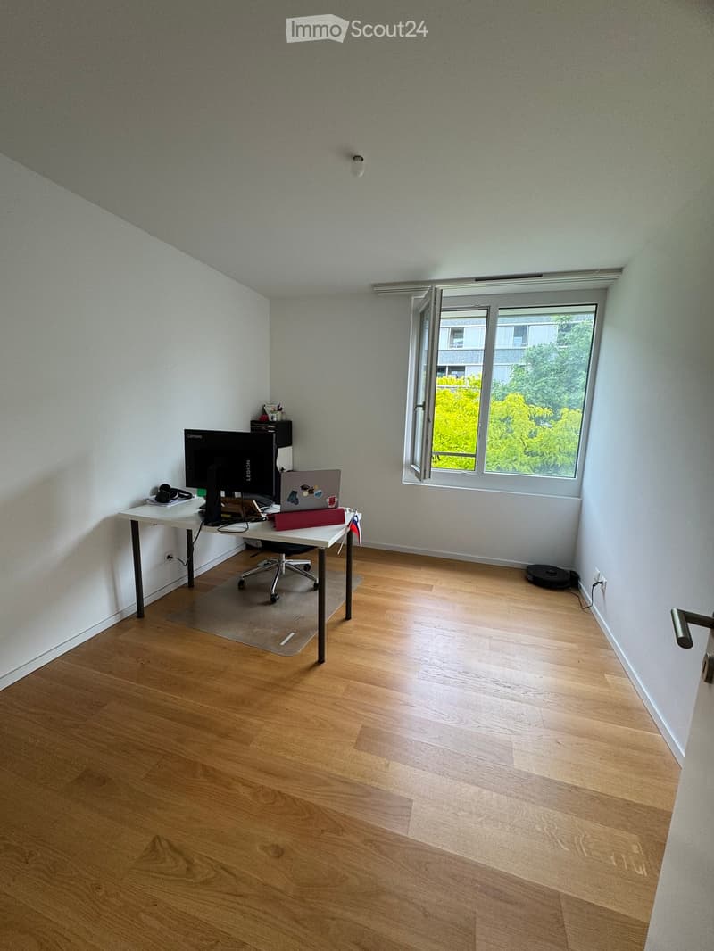 A modern, well-lit room with wooden flooring, a large window, a desk with a laptop, monitor, and chair, and a small shelf with office supplies.