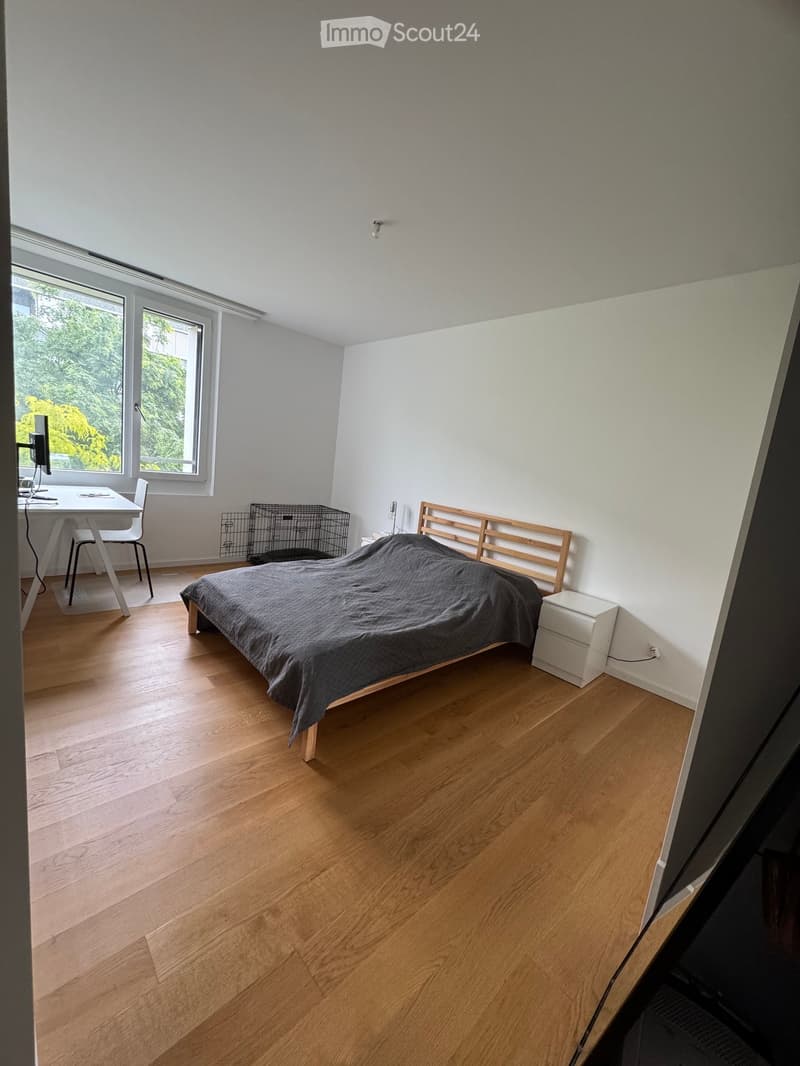 Modern bedroom with wooden flooring, large windows, a bed with a grey duvet, a small table with a chair, and a screen on a stand.