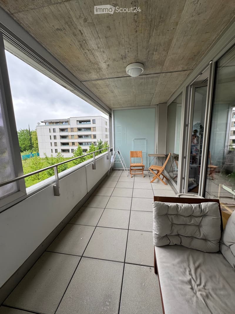 Modern balcony with tiled floor, grey sofa, wooden chairs, small table, and railing overlooking a green area.