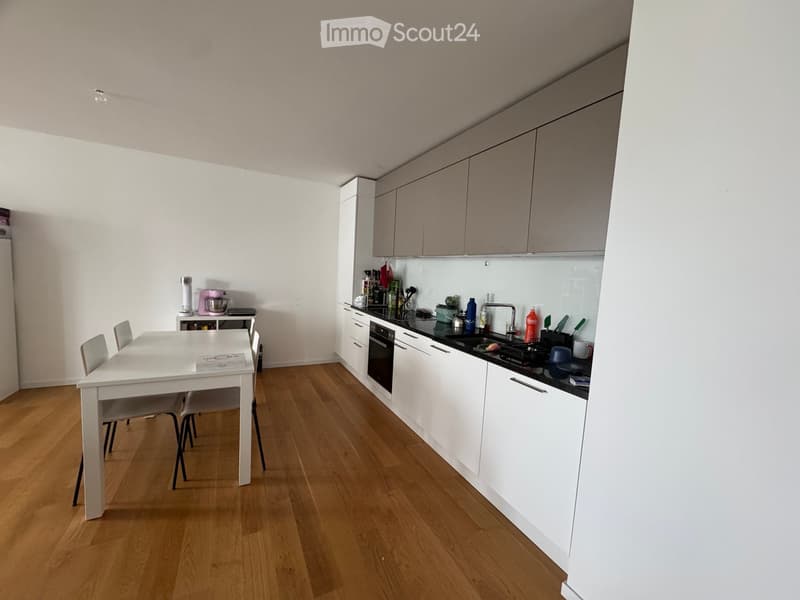 Modern kitchen with white cabinets, wooden flooring, a dining table with chairs, and various appliances including a blender, mixer, and oven.