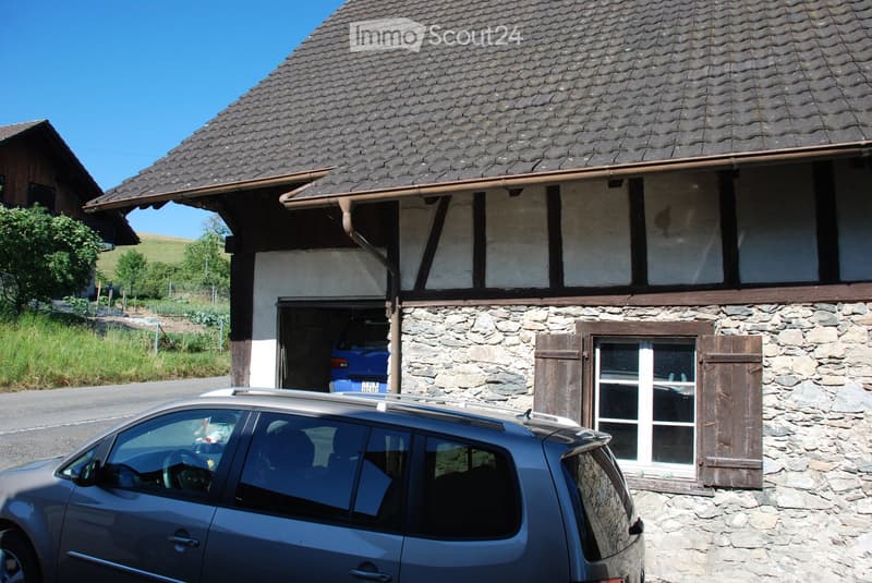 Traditional stone house with a tiled roof, wooden shutters, and a garage. A blue car is parked in the garage, and another car is parked in front of the house.