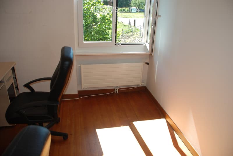 A small, unfurnished room with wooden flooring, a large window with a view of greenery, a black office chair, a radiator, and a small desk.