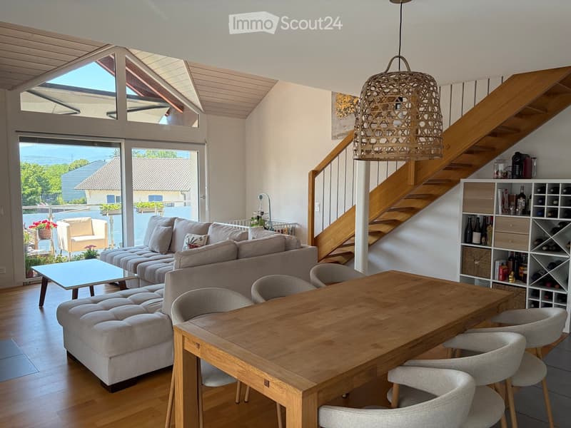 Bright living and dining area with a grey sectional sofa, ottoman, coffee table, dining table with chairs, and a wine rack. The space features wooden flooring, a staircase with a hanging pendant light, and large windows offering a scenic view.