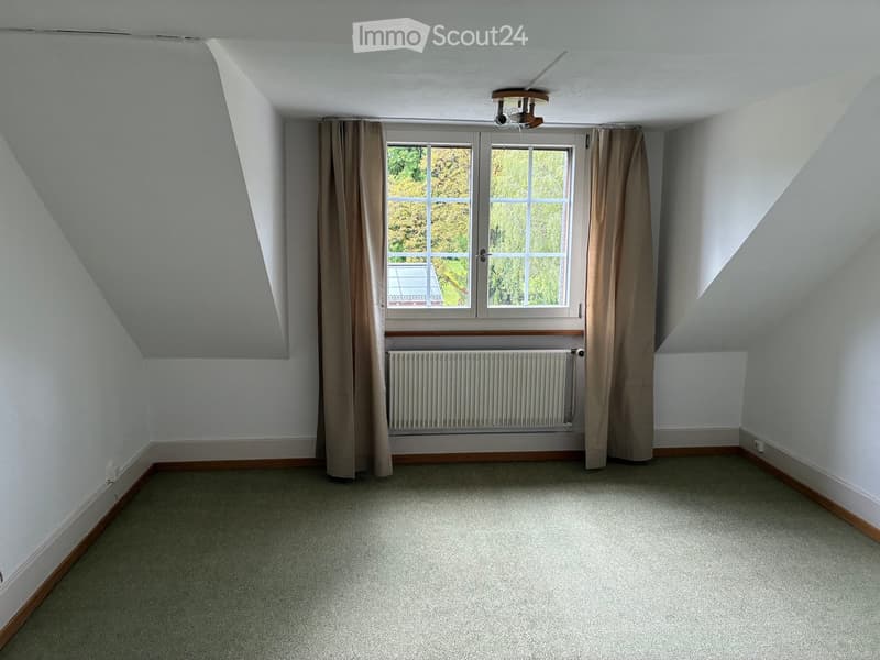 Empty attic room with sloped ceilings, large window with curtains, radiator, and carpeted floor.