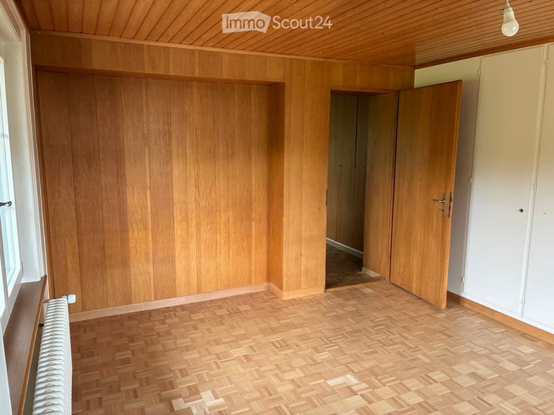 Empty room with wooden walls, herringbone wooden flooring, and a radiator. Features a large window and an open doorway leading to another room.