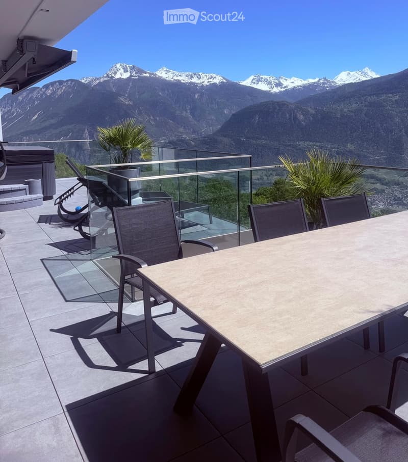 Modern balcony with a glass railing, overlooking a mountain range. Features a dining table with chairs, sun loungers, and potted plants.