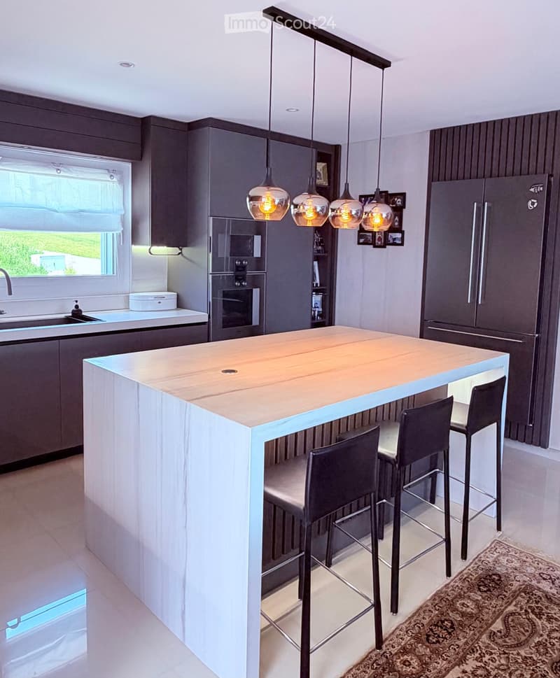 Modern kitchen with a central island, bar stools, pendant lights, and built-in appliances including a refrigerator and oven. The island features a light wood countertop and white base. A window with blinds allows natural light to enter.