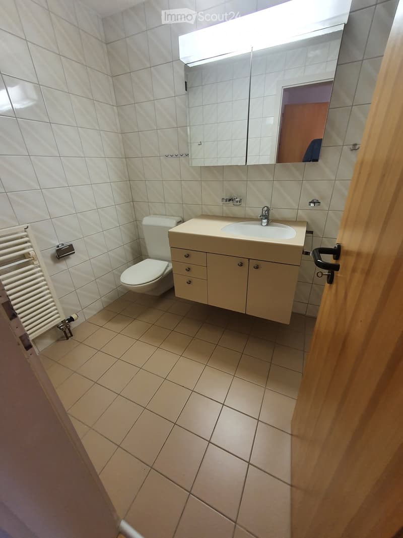 Modern bathroom with white tiled walls, beige tiled floor, a toilet, a sink with a cabinet, a mirror, and a radiator.