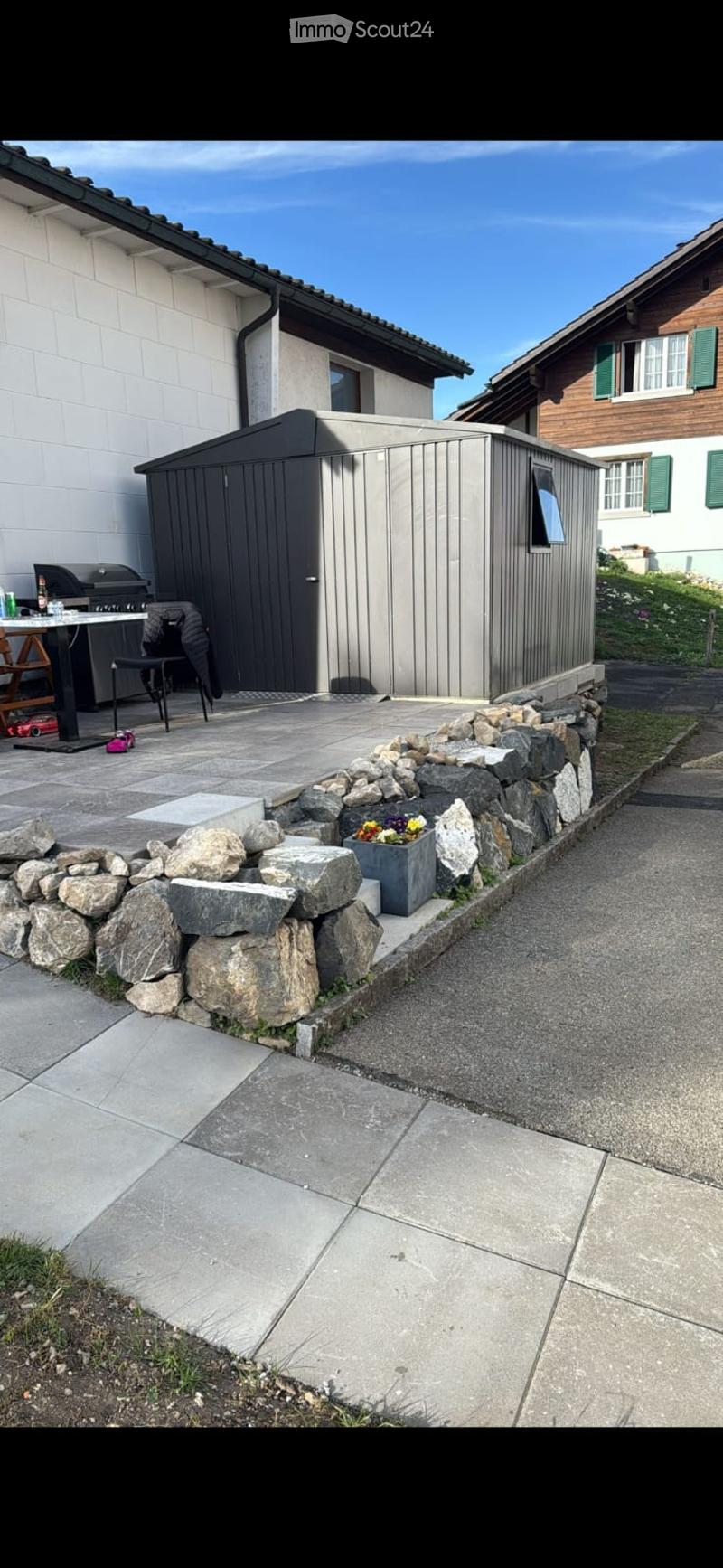 Outdoor patio area with a stone border, a small shed, a grill, a table, and a flower pot.