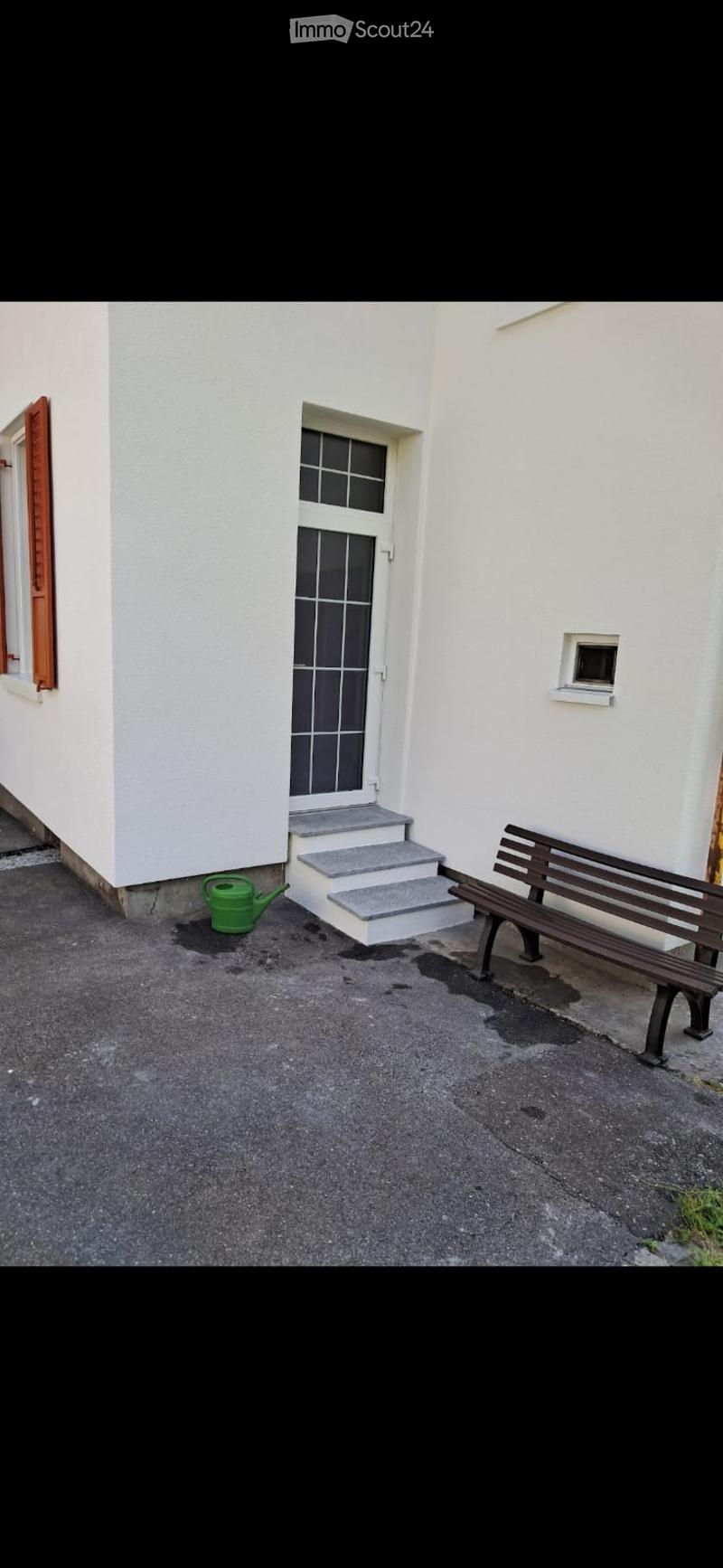 A small outdoor area with a white building entrance, a wooden bench, and a green watering can on the ground.