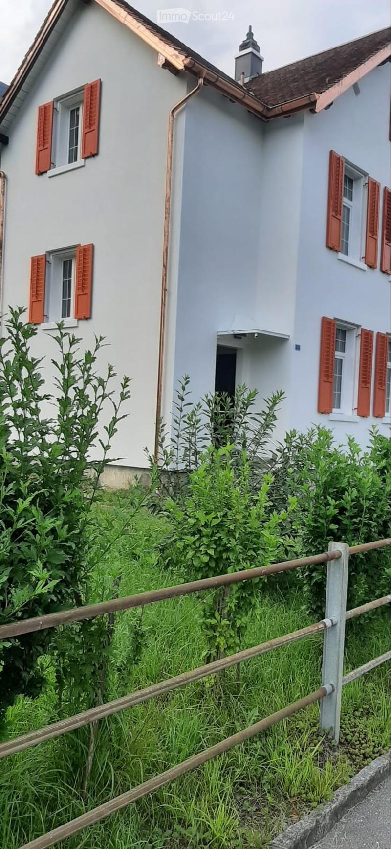 A two-story house with light blue walls, red shutters, and a copper roof. The house has multiple windows and a small balcony on the upper floor. The front yard is enclosed by a wooden fence and is overgrown with greenery.