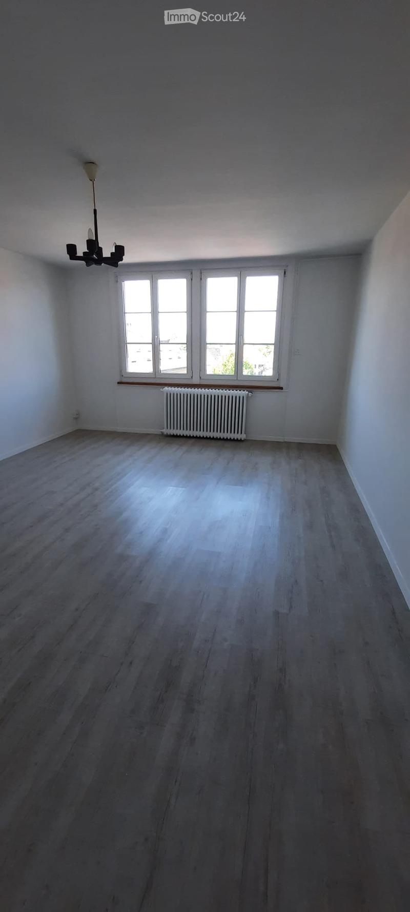 Empty room with wooden flooring, white walls, large windows, radiator, and a chandelier.