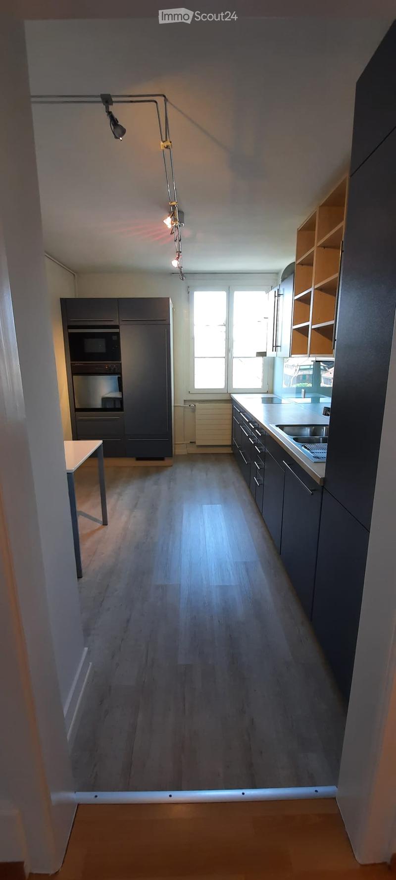 Modern kitchen with dark grey cabinets, stainless steel appliances, wooden flooring, and a small table near the entrance.