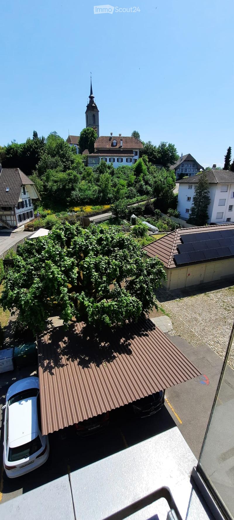 Aerial view of a residential area with multiple houses, trees, and a church tower in the background. A car is parked under a metal-roofed structure, and there is a garage with solar panels on the roof.
