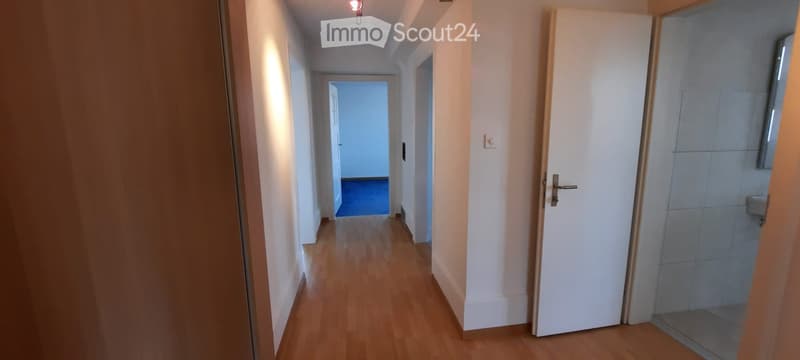 A clean, well-lit hallway with wooden flooring, white walls, and several doors leading to different rooms.