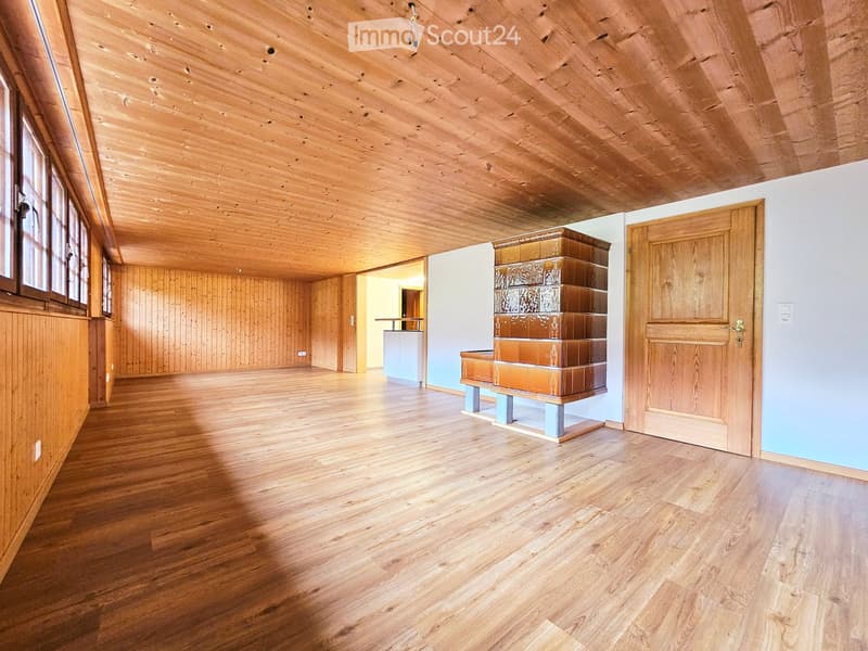 Empty room with wooden floors, wooden walls, and a wooden ceiling. Features a large window, a door, and a small piece of furniture in the background.