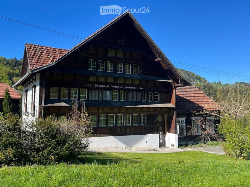 Traditional two-story wooden house with a steeply pitched roof, multiple windows, and a covered balcony area. The house features a mix of white and dark wood siding, with a well-maintained lawn and surrounding greenery.