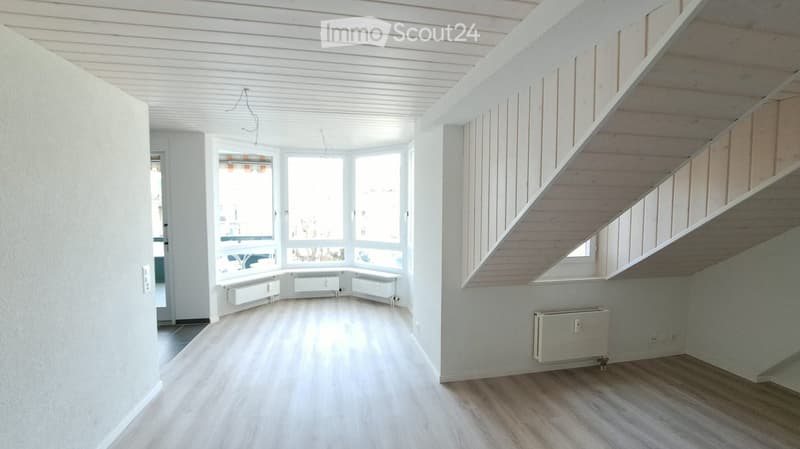 Bright, empty room with wooden flooring, white walls, large windows, and a staircase leading upwards. No furniture or personal items present.