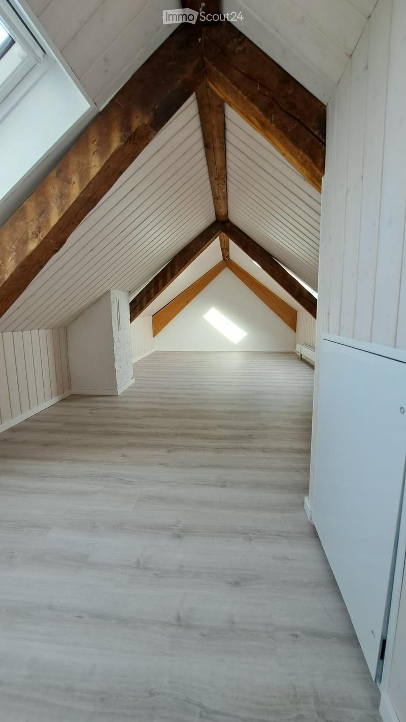 Empty attic space with wooden beams, slanted ceilings, and natural light from skylights. The floor is light grey concrete, and the walls are white with exposed wooden beams.