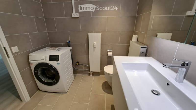 Modern bathroom with a white sink, chrome faucet, toilet, and a compact washing machine. The walls are tiled in a neutral color scheme, and a large mirror is mounted above the sink.