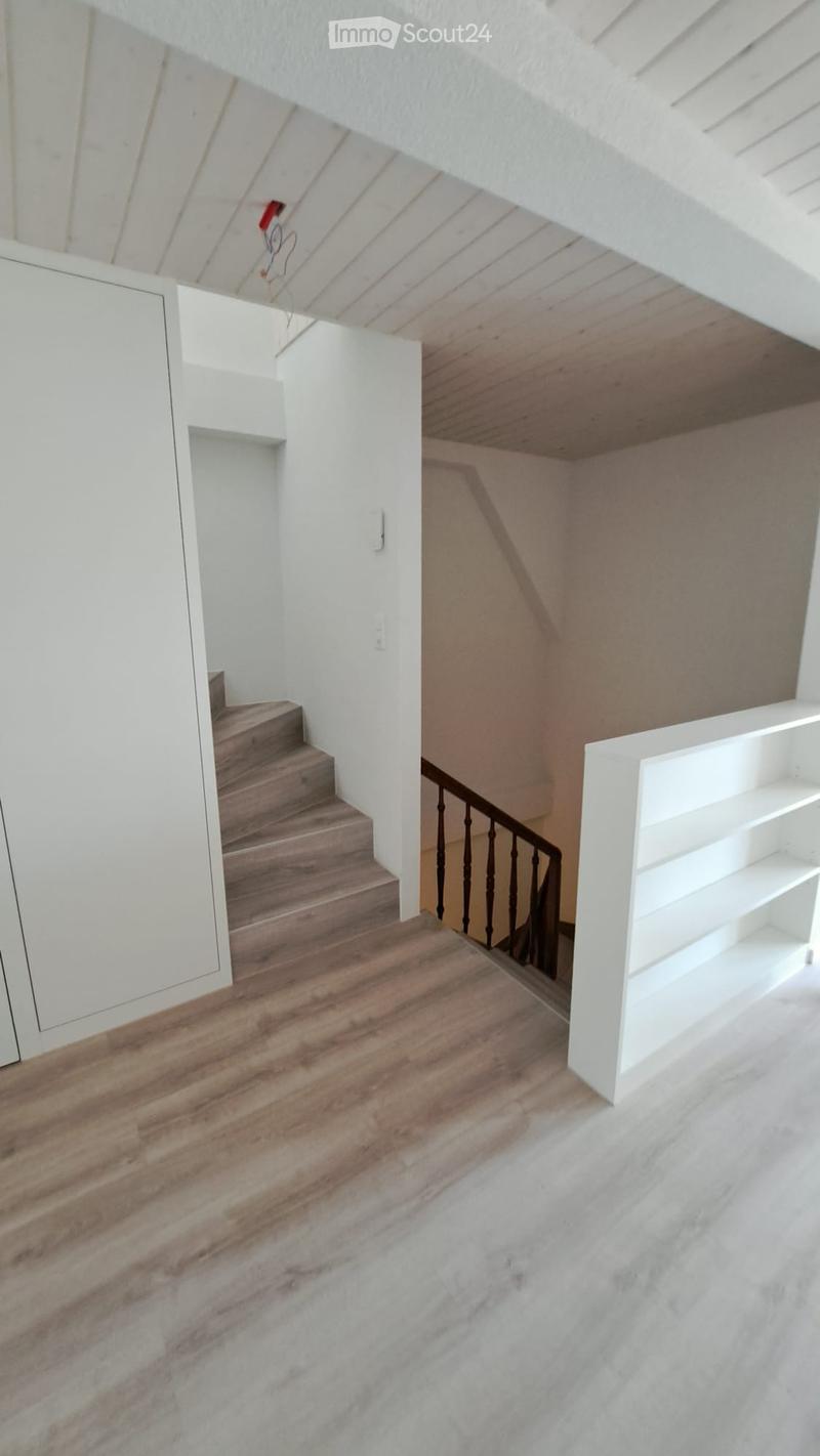 Modern hallway with wooden flooring, white walls, and a staircase leading upwards. Features a white built-in shelf on the right side and a dark wooden railing on the staircase.
