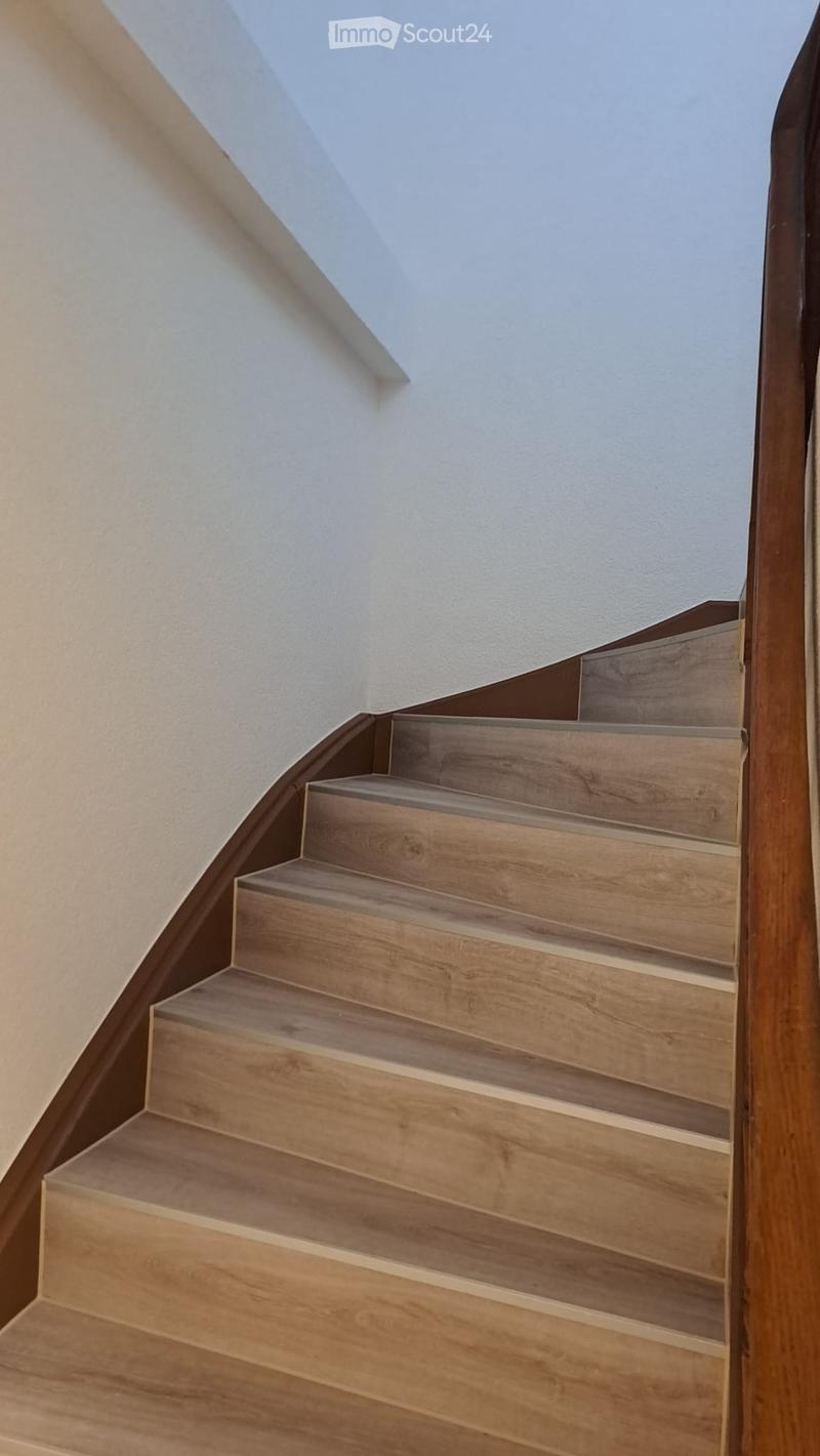 Wooden staircase with a modern design, featuring light-colored steps and a dark wooden handrail, set against white walls and a slanted ceiling.