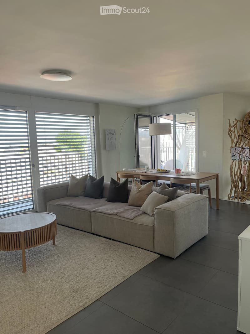 Modern living room with grey sectional sofa, coffee table, dining table, and floor lamp. Large windows with blinds and access to a balcony.