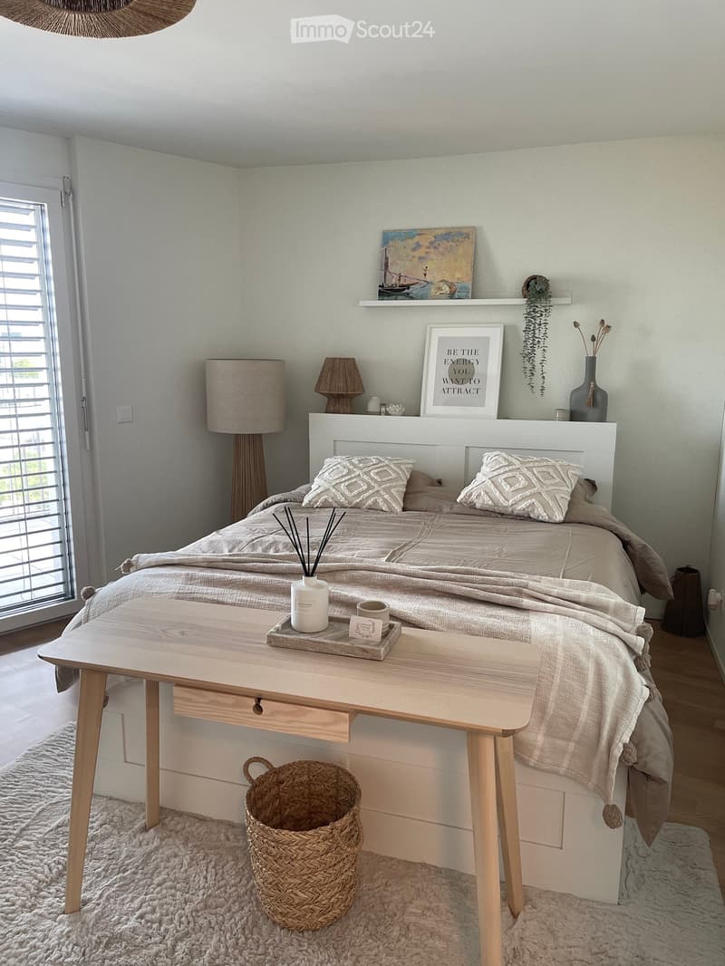 A modern, well-lit bedroom with a large bed, nightstands, a wooden table, and decorative items including a vase, picture frame, and a tray with a coffee mug.