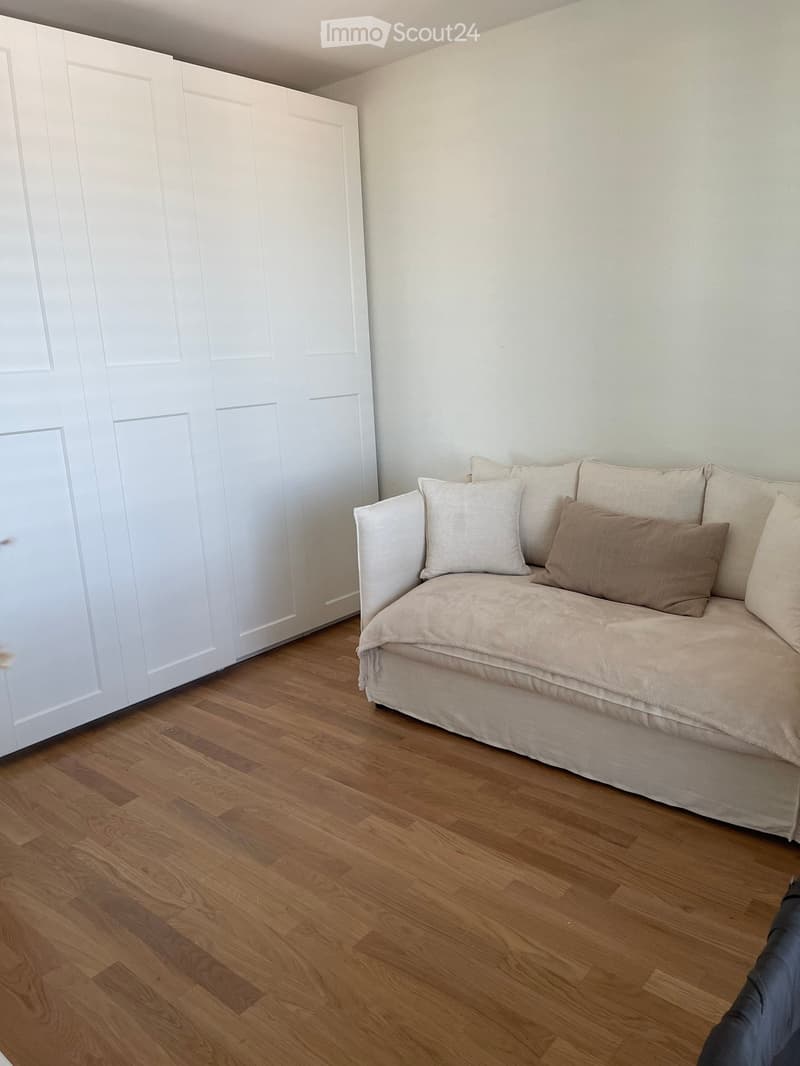 A small, cozy living space with a wooden floor, a white door, and a beige sofa with multiple pillows.