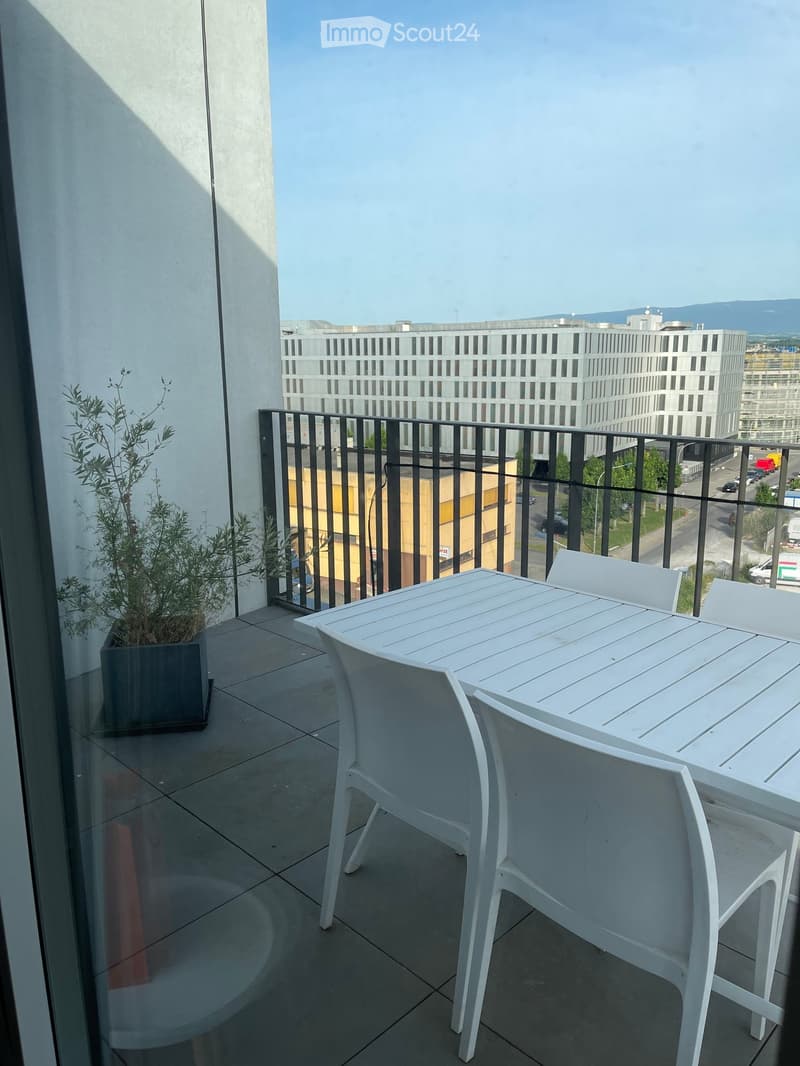 Balcony with a white table and chairs, a potted plant, and a view of a modern building and cityscape.