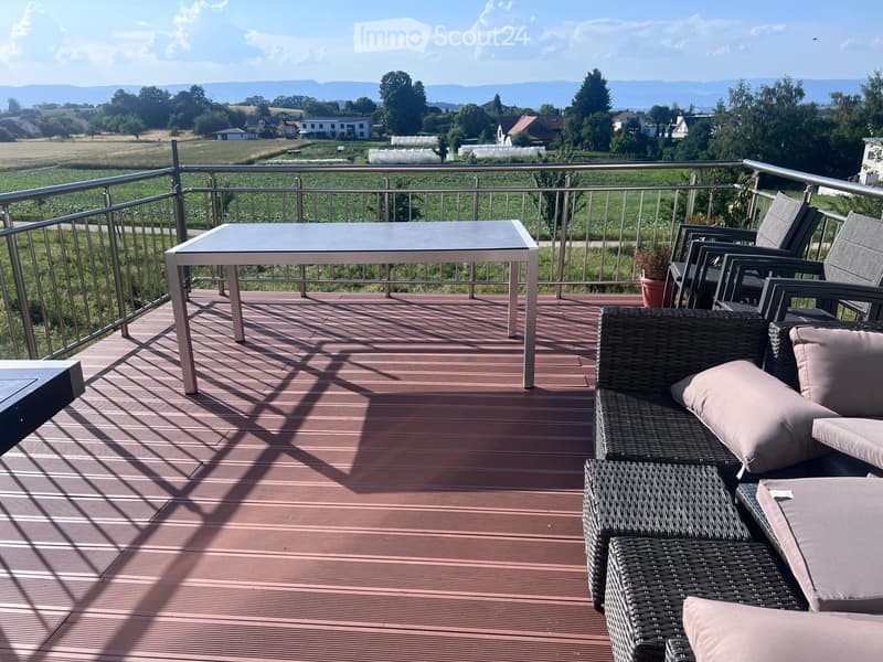 Balcony with wooden decking, metal railing, and outdoor furniture including a table, chairs, and a wicker sofa with cushions.