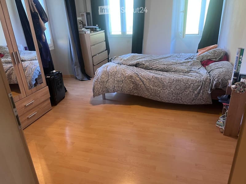 Bedroom with wooden flooring, a double bed with patterned bedding, a wardrobe, a nightstand, and a mirror. A suitcase is placed near the wardrobe.