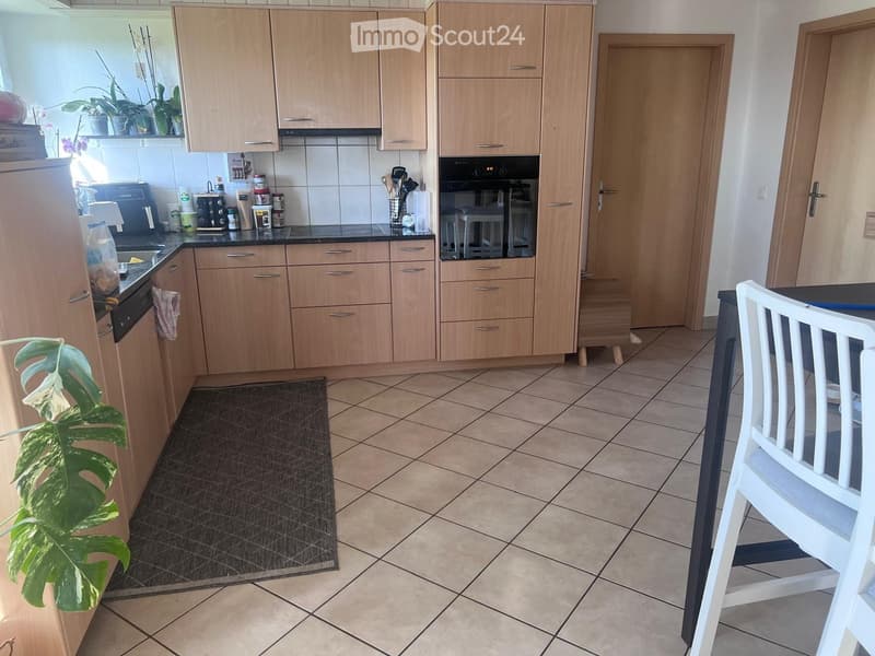 Modern kitchen with light wooden cabinets, tiled floor, and various appliances including a refrigerator, oven, and dishwasher. A small dining area with a white chair and table is visible, along with a potted plant near the counter.