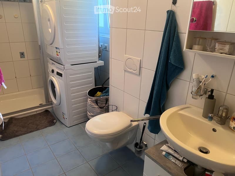 A modern bathroom with a toilet, sink, washing machine, and a shower area. The sink has a toothbrush holder and toothpaste, and a towel hangs on the wall. A mirror is above the sink, and a small trash bin is near the washing machine.