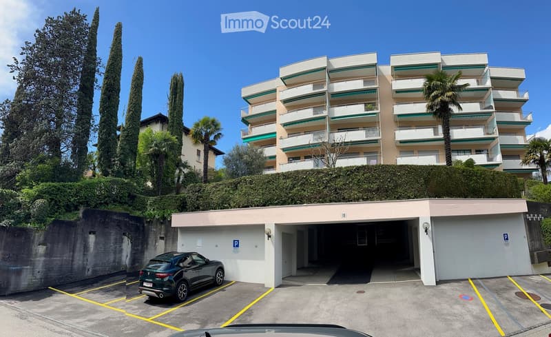 Modern multi-story apartment building with balconies and green awnings, surrounded by tall trees and a hedge. A parking garage is visible in the foreground with a parked car.