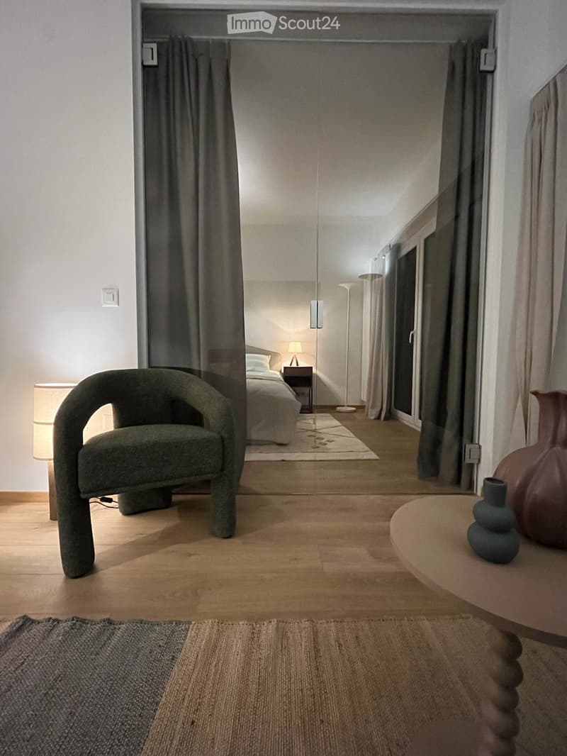 Modern living space with a dark green armchair, round wooden table with vases, and a visible bedroom area with a bed and nightstand.