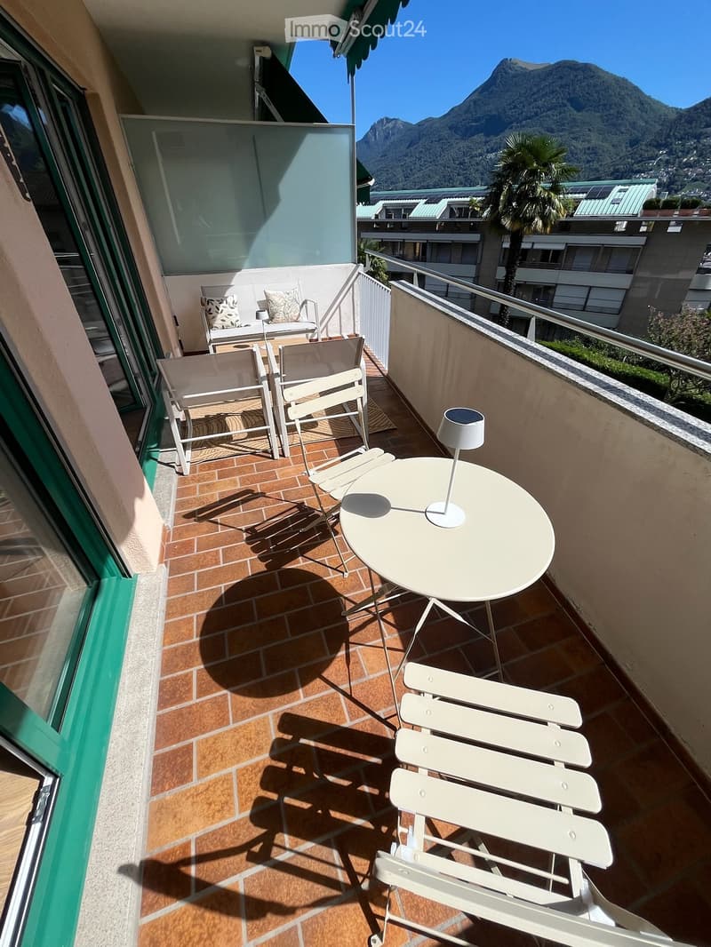 Balcony with tiled floor, white table, chairs, and a lamp, overlooking a scenic mountain view.