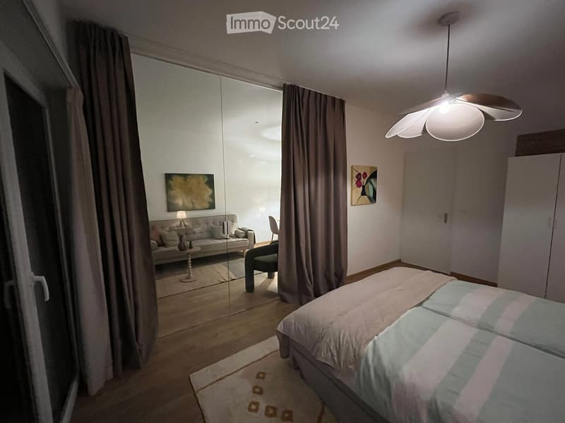 Modern bedroom with a large bed, a sofa, a small table, a chair, a hanging lamp, a mirror, and artwork on the walls.