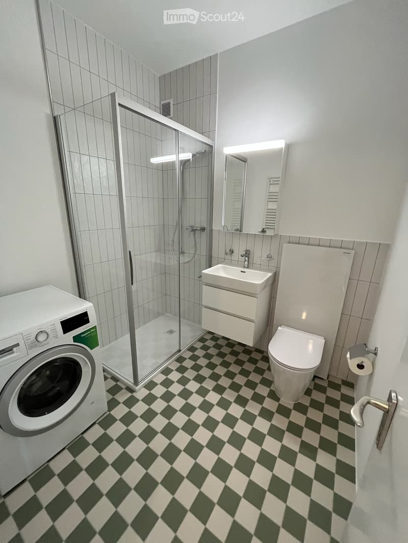Modern bathroom with green and white checkered floor, white toilet, sink with mirror, glass-enclosed shower, and washing machine.