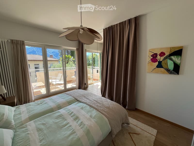 Bright bedroom with two single beds, a nightstand with a lamp, a large painting on the wall, and large windows leading to a balcony with outdoor furniture.