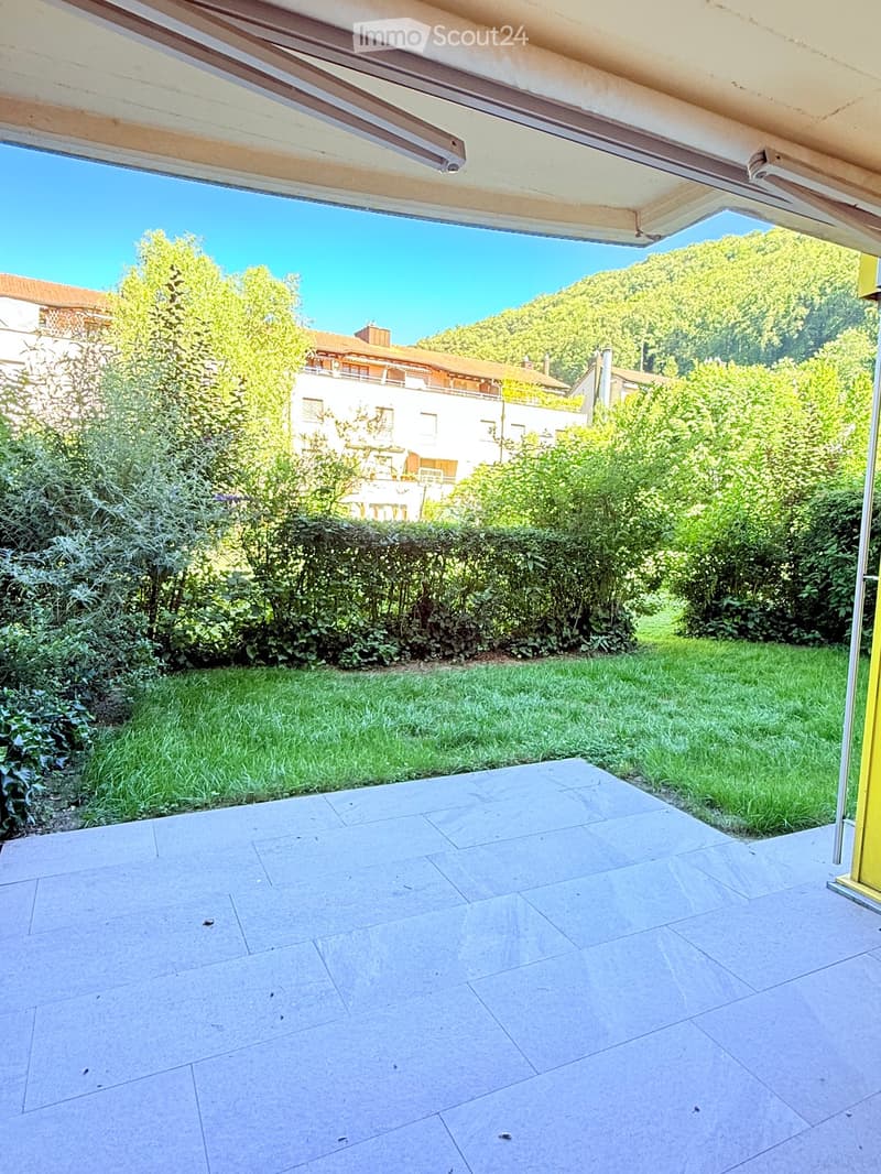 A spacious outdoor patio with large stone tiles, surrounded by green grass and bushes, with a view of a hillside and neighboring houses.