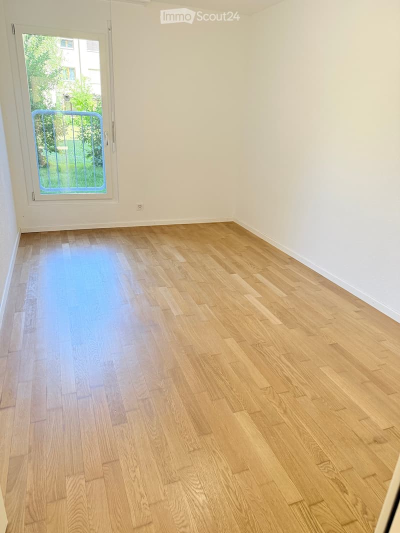 Empty room with light wooden flooring and white walls, featuring a large window with a blue security gate leading outside.