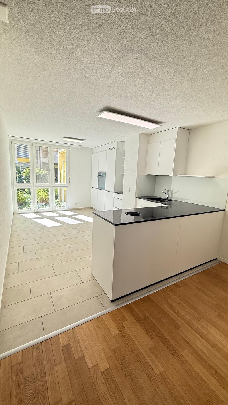 Empty kitchen with white cabinets, a black countertop, and a sink. The floor is a combination of tiled and wooden surfaces, and there is a large window with blinds letting in natural light.